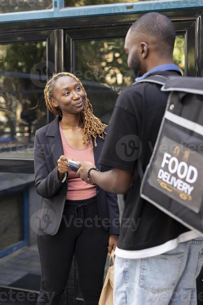 Black woman paying for her takeaway meal order with a contactless card at the doorstep. Delivery man carrying a thermal bag, completes fast food transaction using a wireless POS terminal. photo