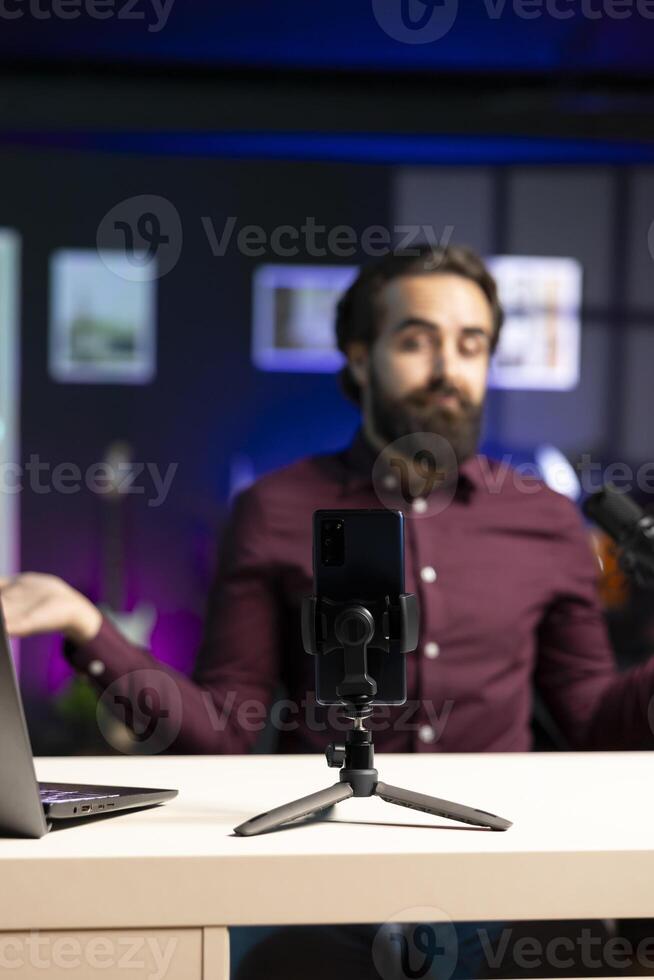Joyful content creator sitting at table talking in front on smartphone on tripod in dimly lit home studio interior, chatting with fans. Man using mobile phone to record vlog in apartment photo