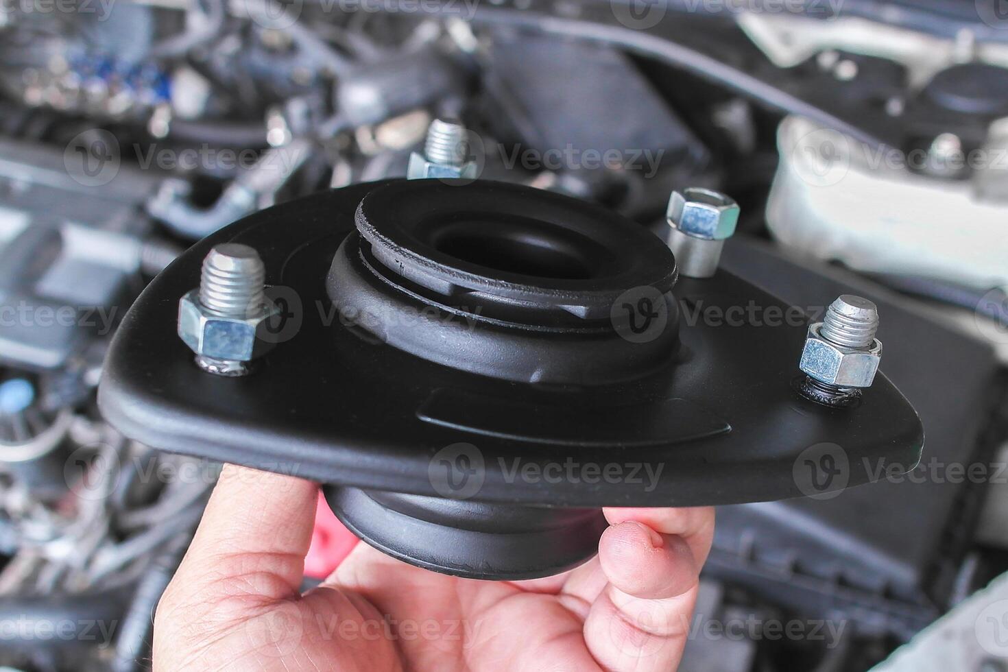 Top Strut Mount, A close-up of hand holds a black automotive strut mount with visible bolts over a car engine. photo