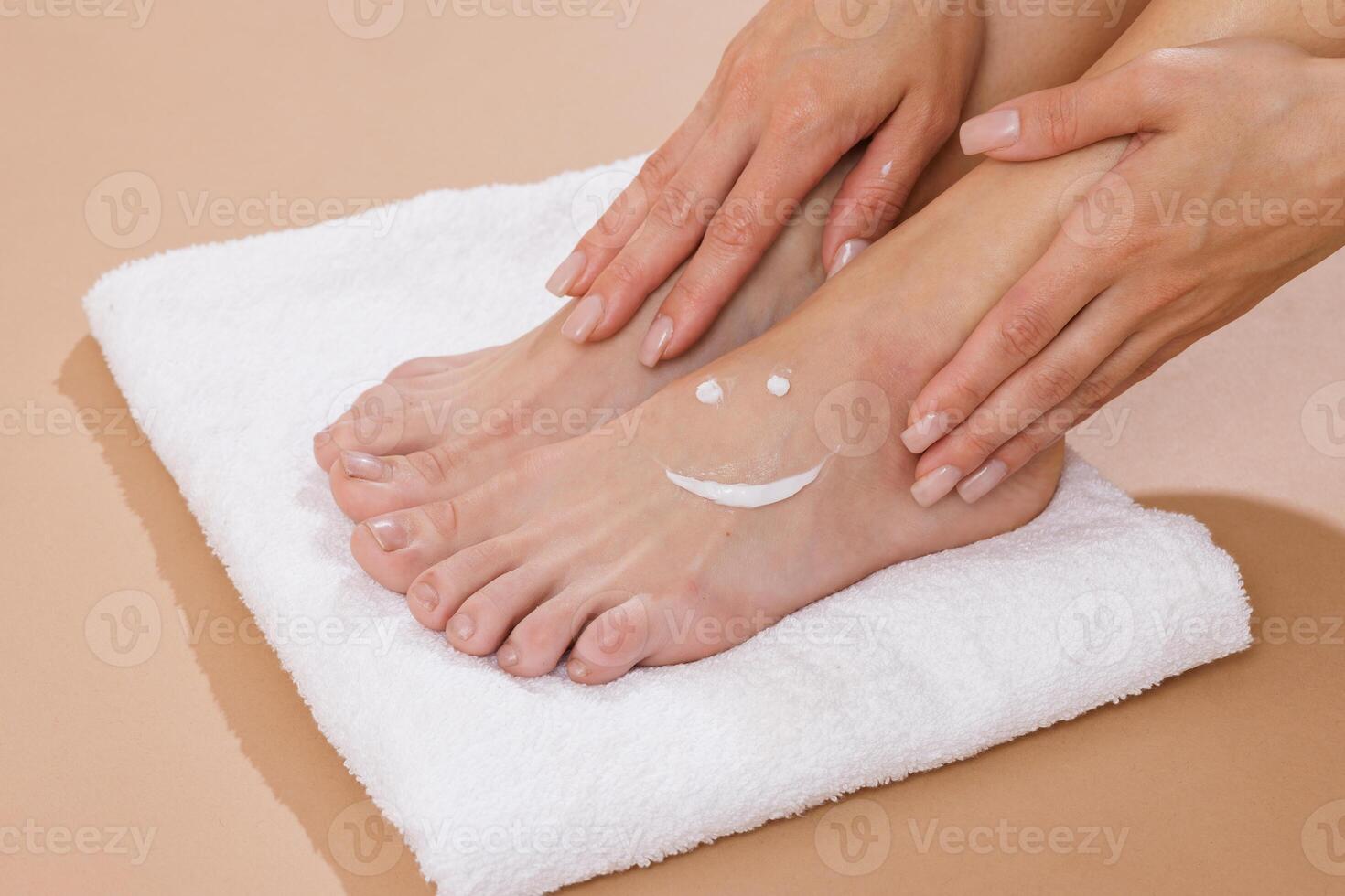 Woman gently rubbing foot cream into her feet, with a smiley made of moisturizer drawn on the skin. Concept of skincare, wellness, and self-care on a white towel background. photo