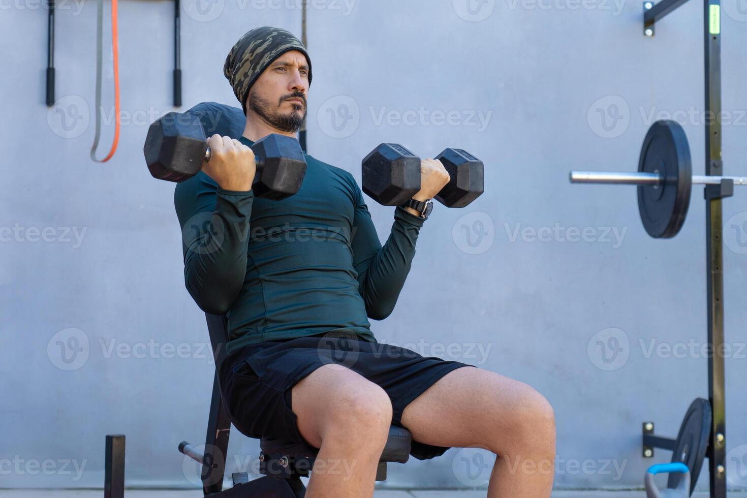 Determined male athlete lifting dumbbells while performing seated bicep curls outdoors photo