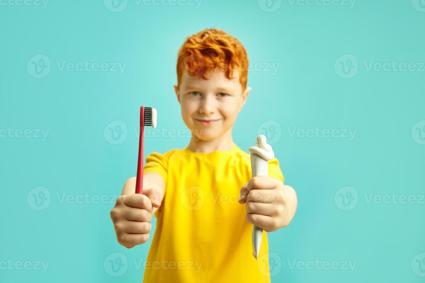 Red haired little boy holding brush tooth and tube of dental paste, preparing to clean teeth, wearing in yellow t shirt standing on blue isolated background. Sharpness on the hands, blurred face. photo