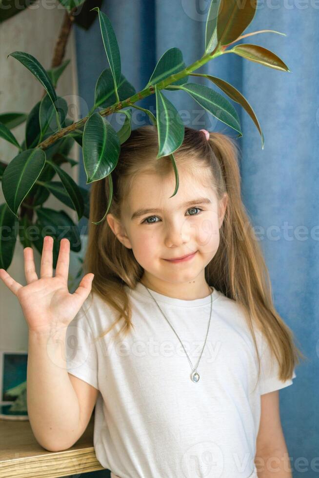 Adorable Little Girl with Brown Hair Waving Hand Surrounded by Green Leaves Indoors. photo