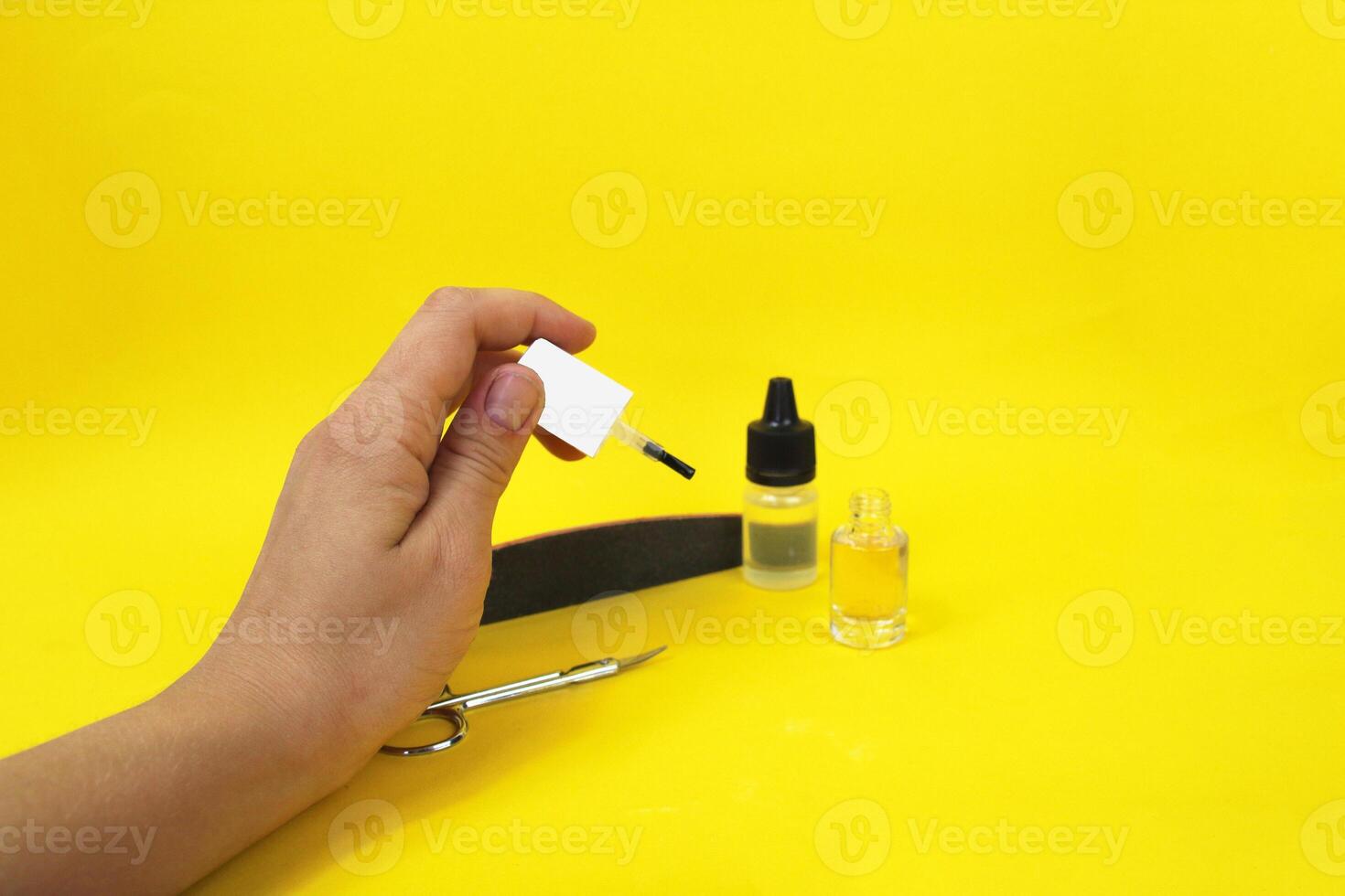Manicure at home. On a yellow background there is cuticle oil and varnish, next to it there are scissors and a nail file. In a woman's hand there is a varnish brush photo