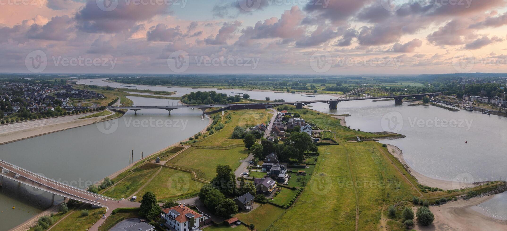 Aerial view of Nijmegen, Netherlands, showcasing the Waal River, De Oversteek, Waalbrug, green fields, residential areas, and scattered clouds in the sky. photo