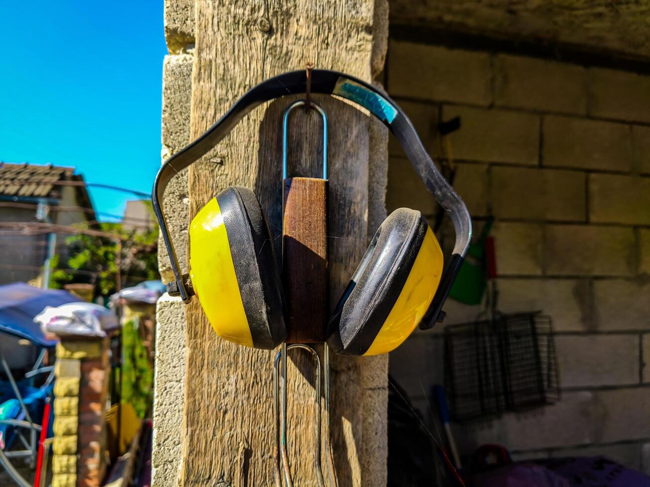 A pair of earplugs, one yellow and one black, are hanging from a wooden post. The earplugs are attached to a metal hook, which is hanging from the post. The post is located in a yard photo