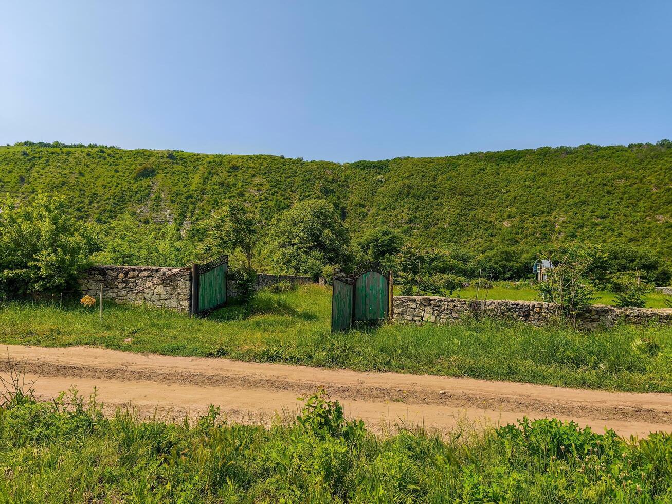 A green hillside with a stone wall and a gate. The gate is open and the road is dirt photo