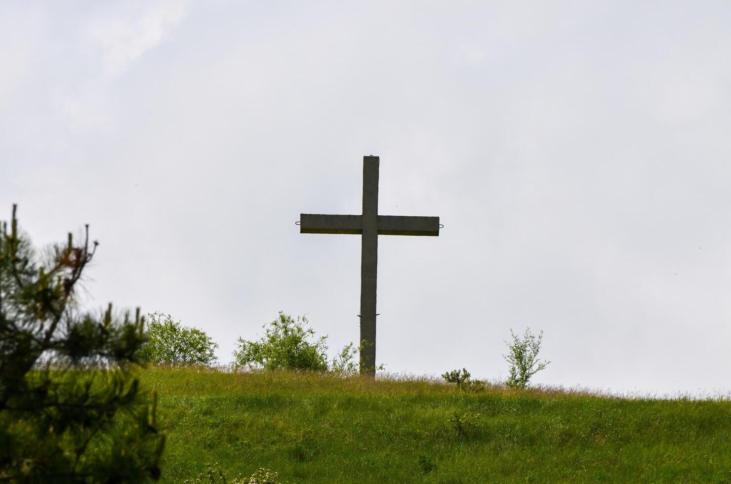A wooden cross is standing on a hillside in a grassy field. The sky is cloudy, and the cross is the only visible object in the scene photo