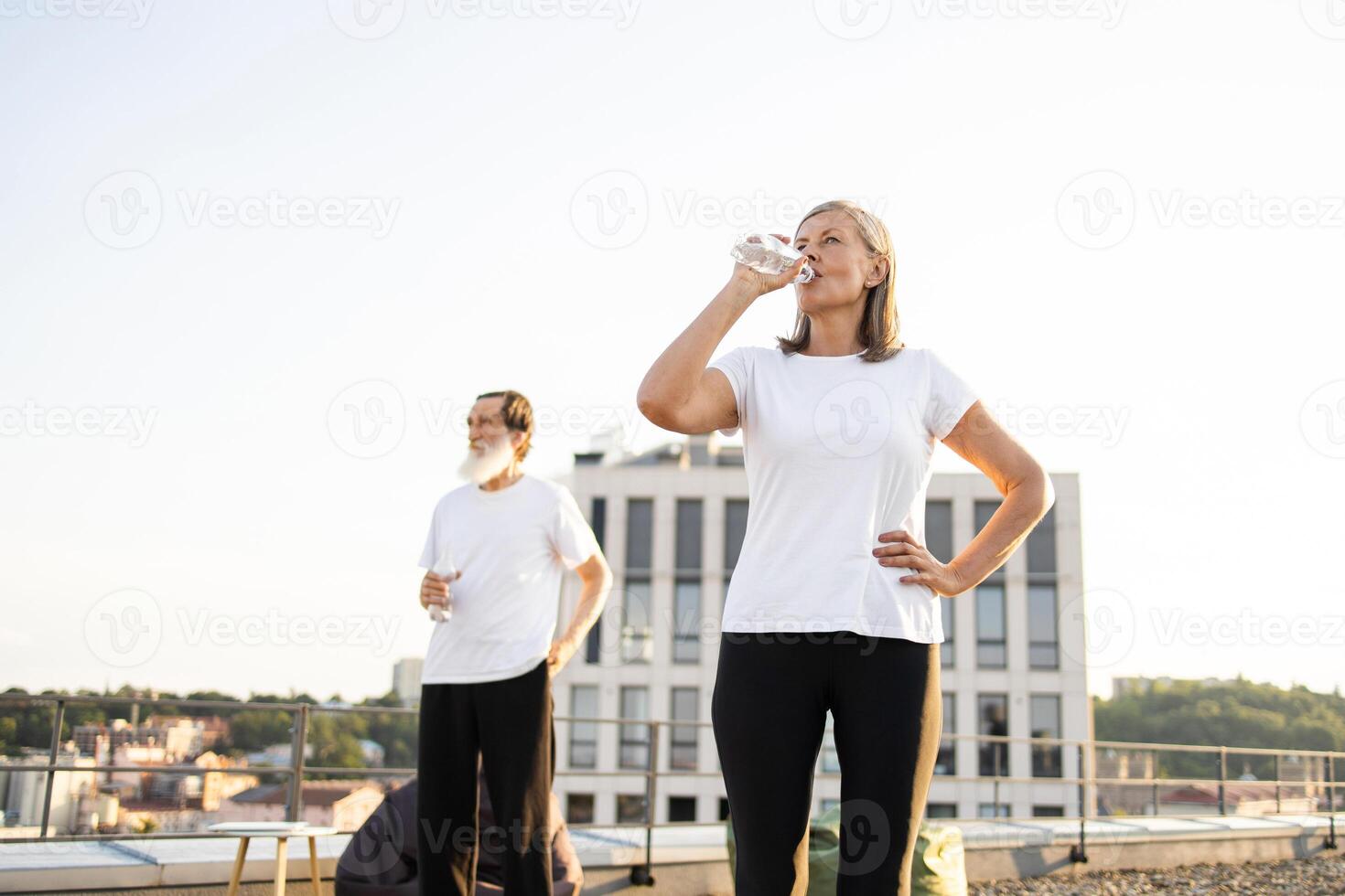 Elderly man and woman in open rooftop setting, enjoying refreshing water after workout outdoors in city. Representing healthy lifestyle, fitness activities, active retirement and responsible hydration photo