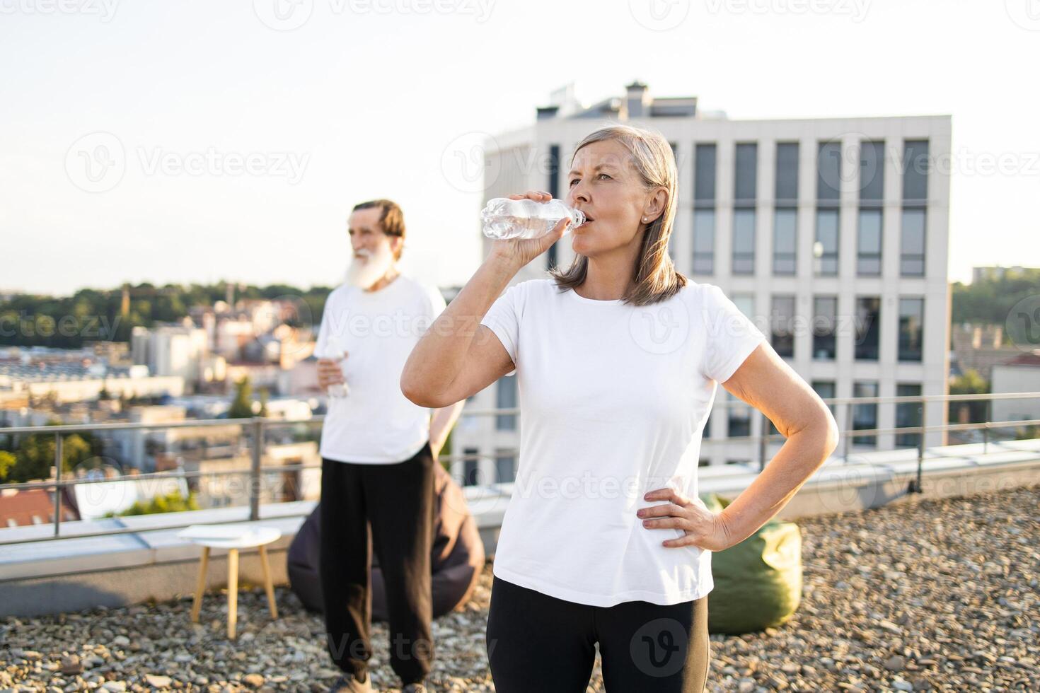 Senior woman drinking water and man standing outside on rooftop, enjoying refreshment. Vibrant composition emphasizing health and fitness. Clear sky and cityscape add energy and inspiration. photo