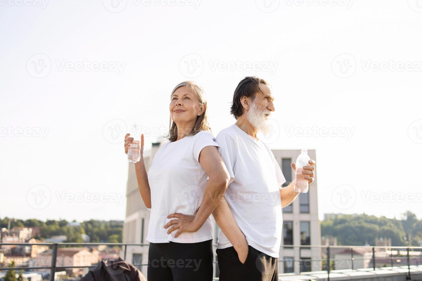Senior couple drinking water on rooftop after workout. Wearing casual activewear, sunny day. Promotes healthy lifestyle, wellness, and hydration. photo