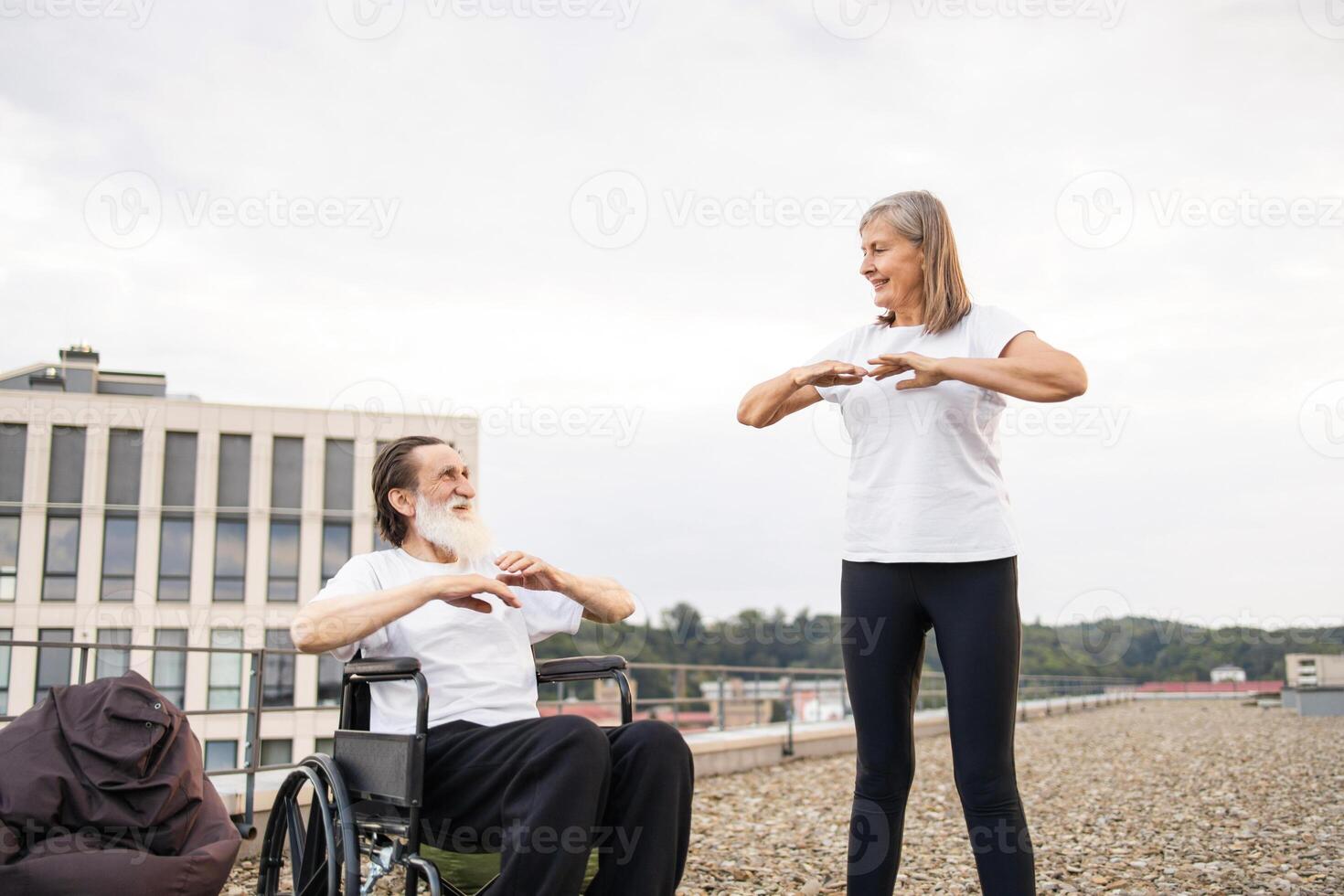Elderly woman assisting man in wheelchair practicing exercises on rooftop, promoting fitness and well-being. Man and woman demonstrating teamwork, inclusivity, and active lifestyle. photo