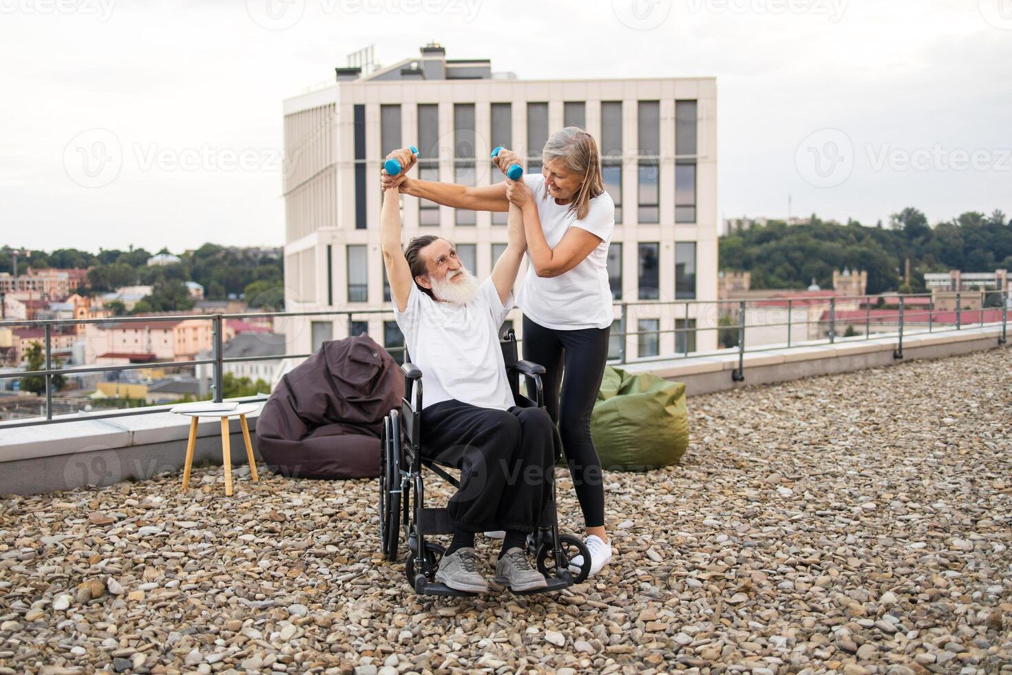 Senior woman assisting wheelchair-bound husband with rehabilitation exercises using dumbbells on rooftop. Bright day enhancing supportive, emotional interaction, emphasizing recovery, togetherness. photo