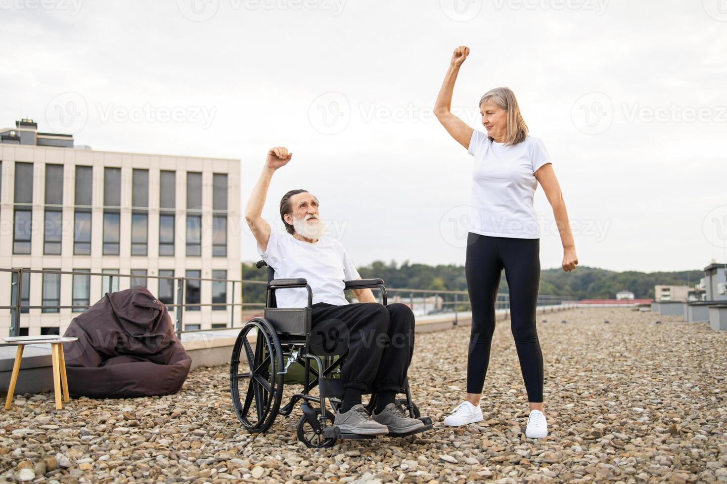 Senior couple, woman stretching, man in wheelchair raising arm outdoors on rooftop. Depicts teamwork, support, active lifestyle, disability awareness, mutual encouragement, outdoor exercise setting. photo