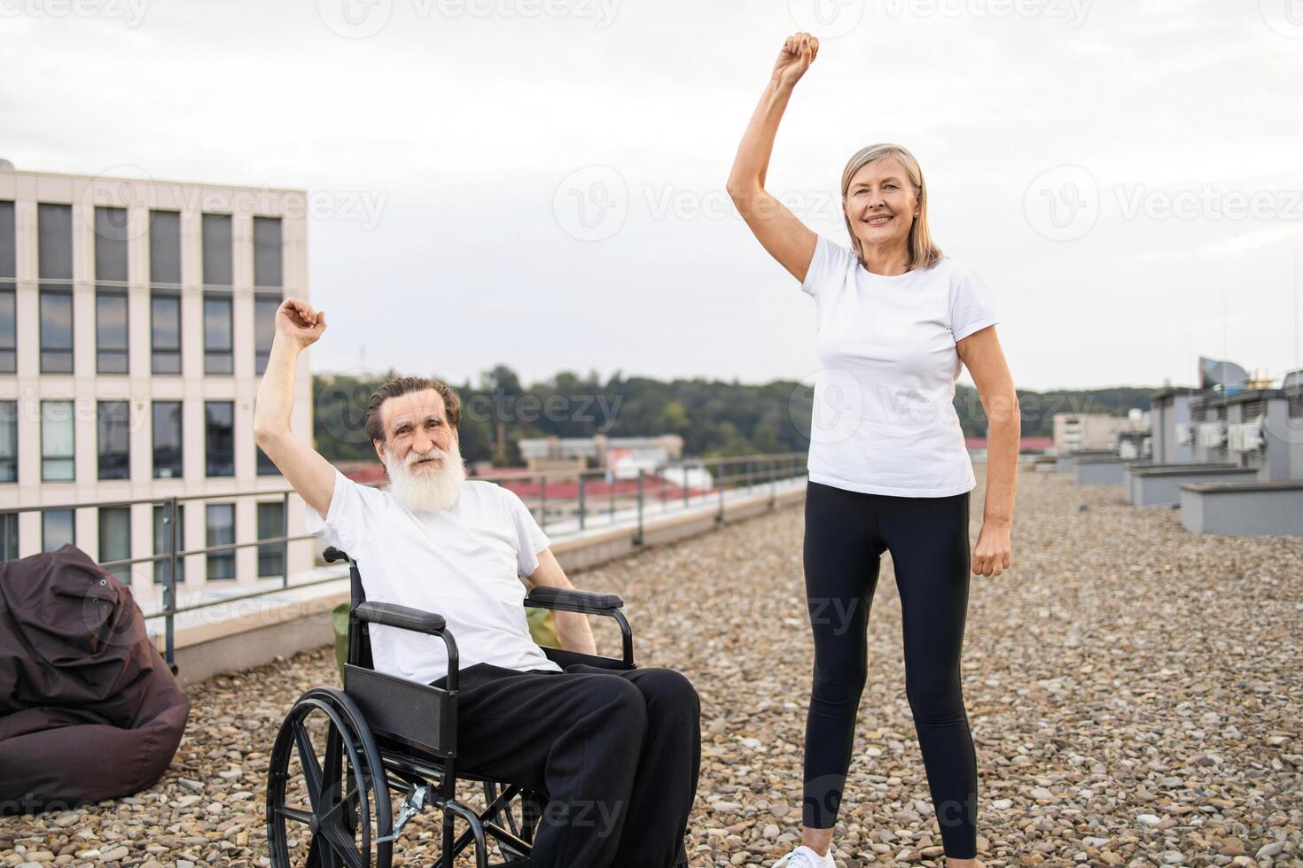 Elderly man in wheelchair and woman standing together lifting arms outside urban rooftop photo