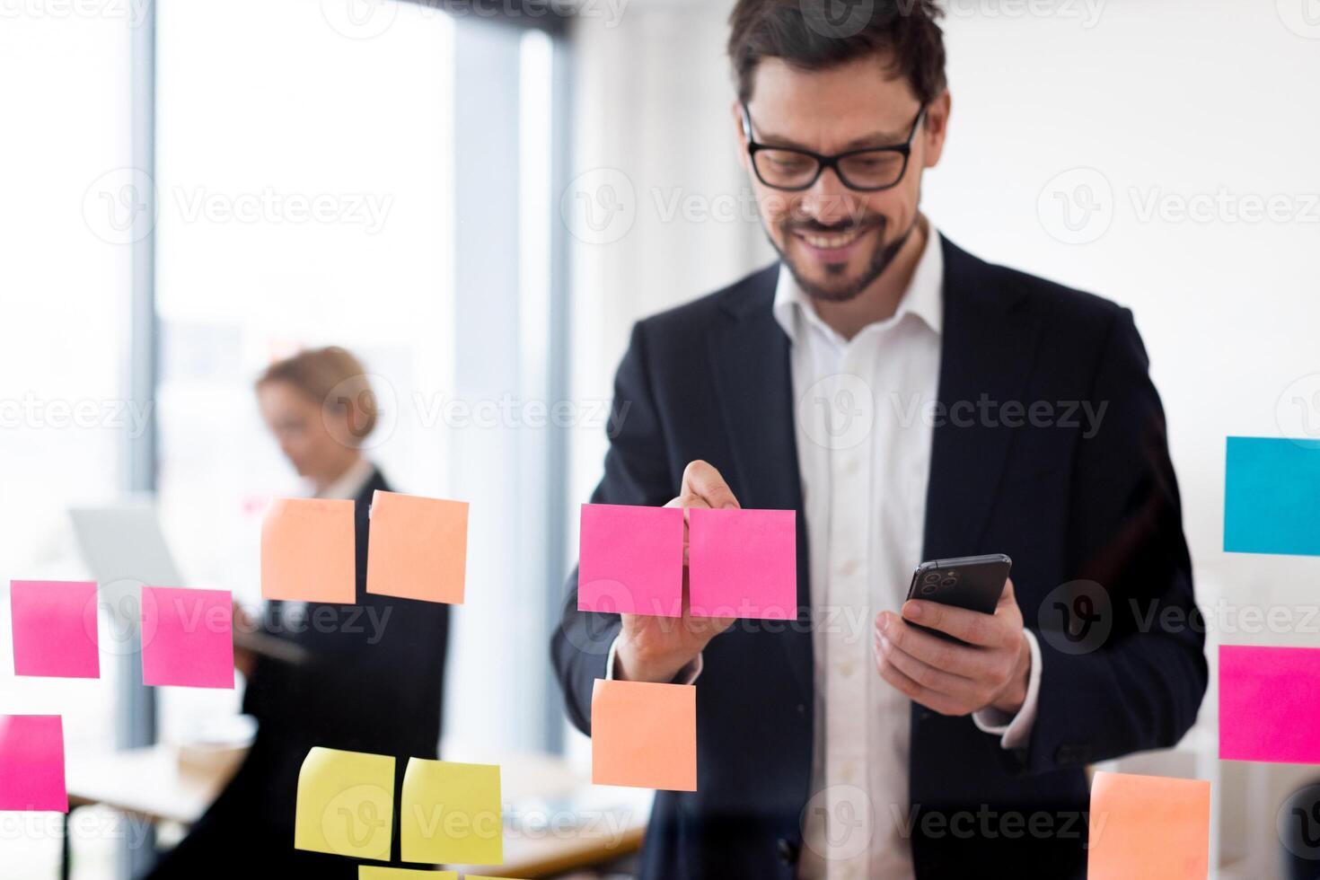 Adult male organizing tasks with colorful sticky notes on glass board, holding smartphone. Office backdrop with modern workplace vibes. photo