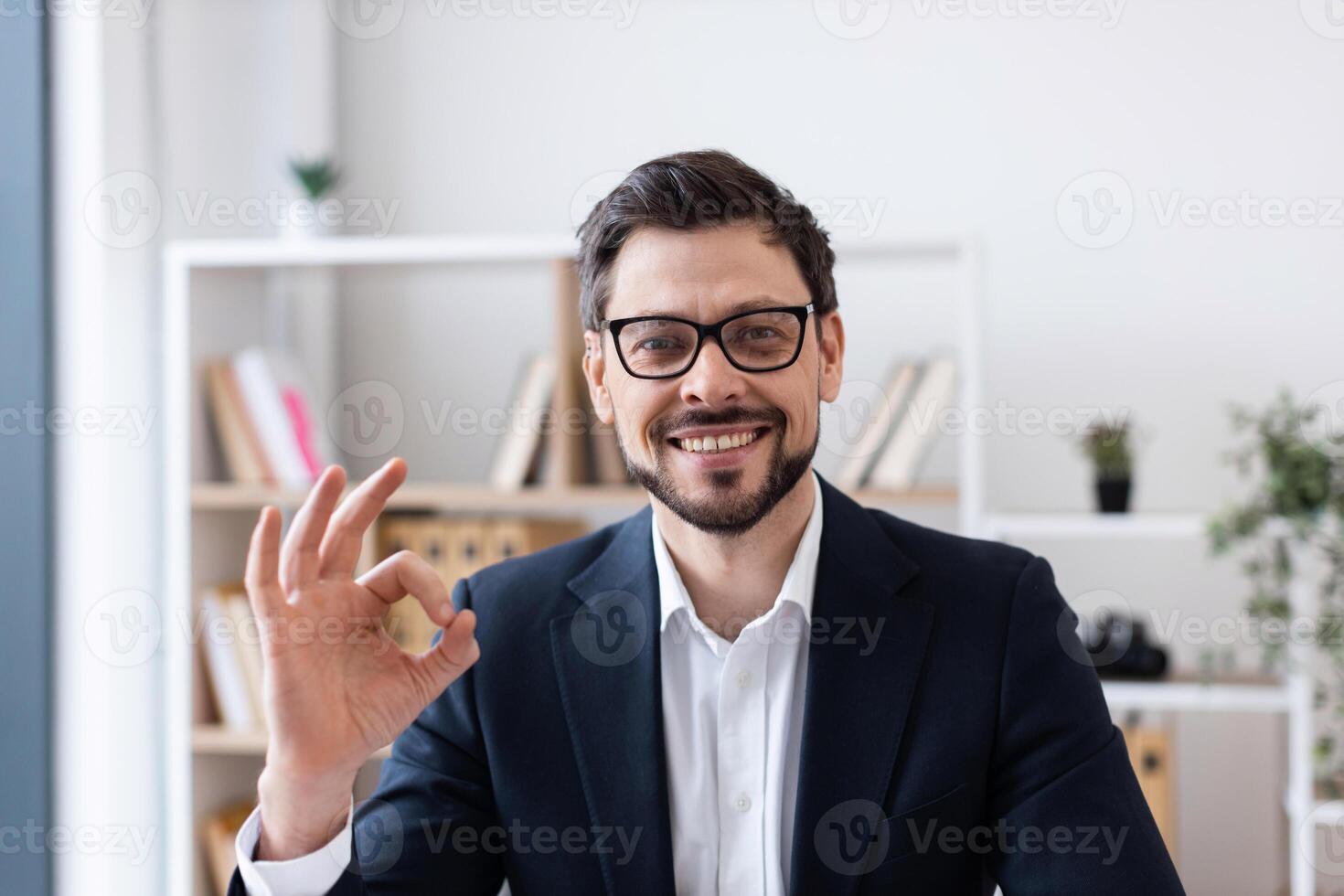 Professional adult man with beard and glasses wearing formal attire smiling in office. Hand gesture expressing agreement or positivity. Background shows shelves with books and decoration items. photo
