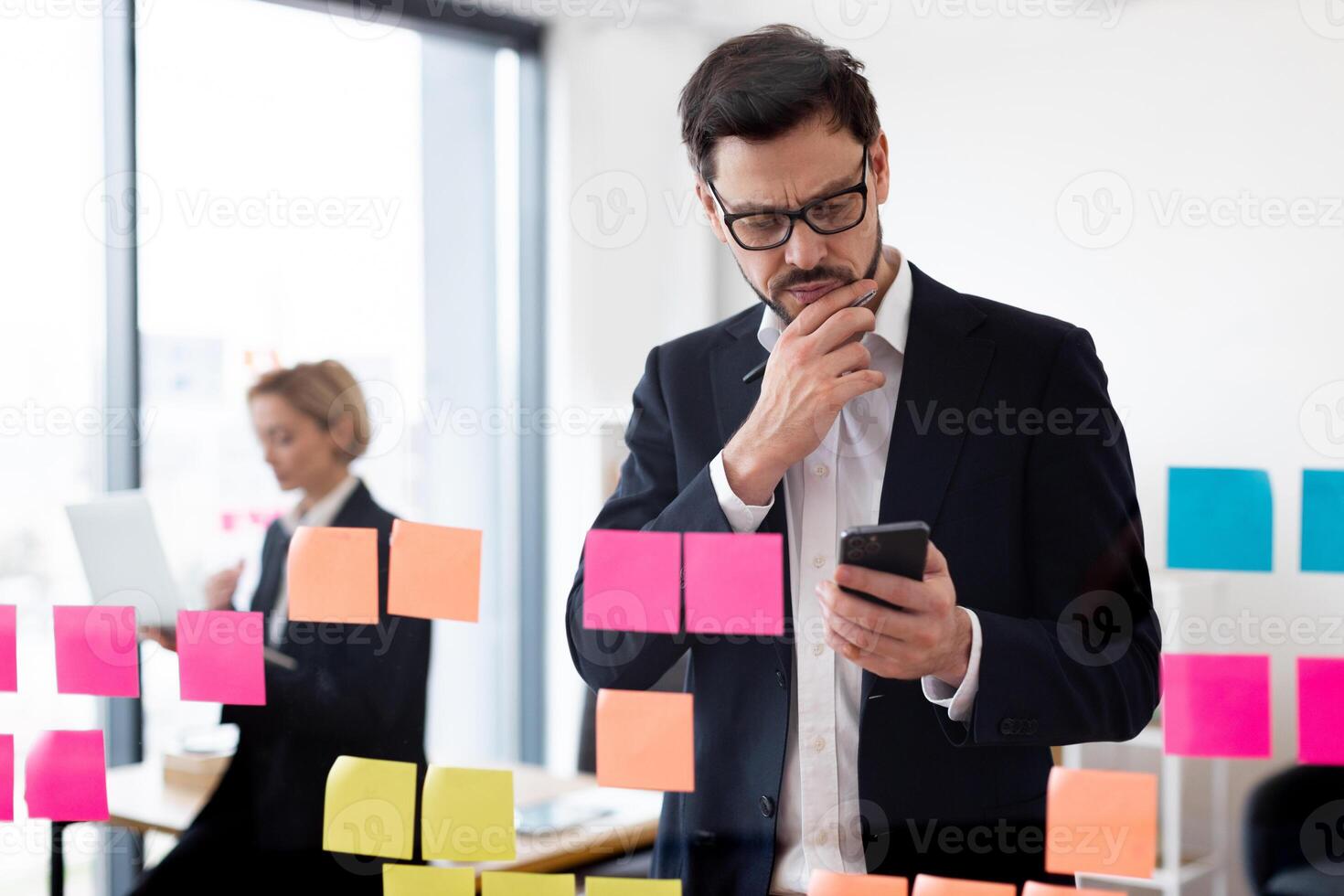 Confident adult man in formal suit analyzing colorful sticky notes on glass in modern workspace. Assisting female colleague visible in background. photo