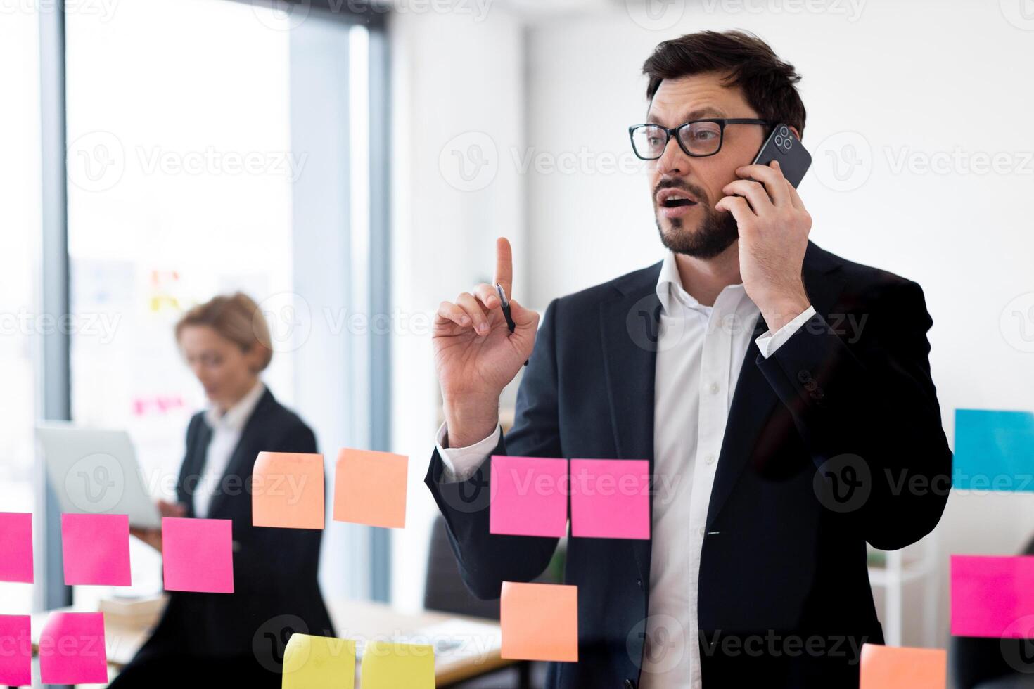 Confident adult businessman discussing ideas on phone while collaborating with female colleague in modern office setting with organized transparent glass board photo