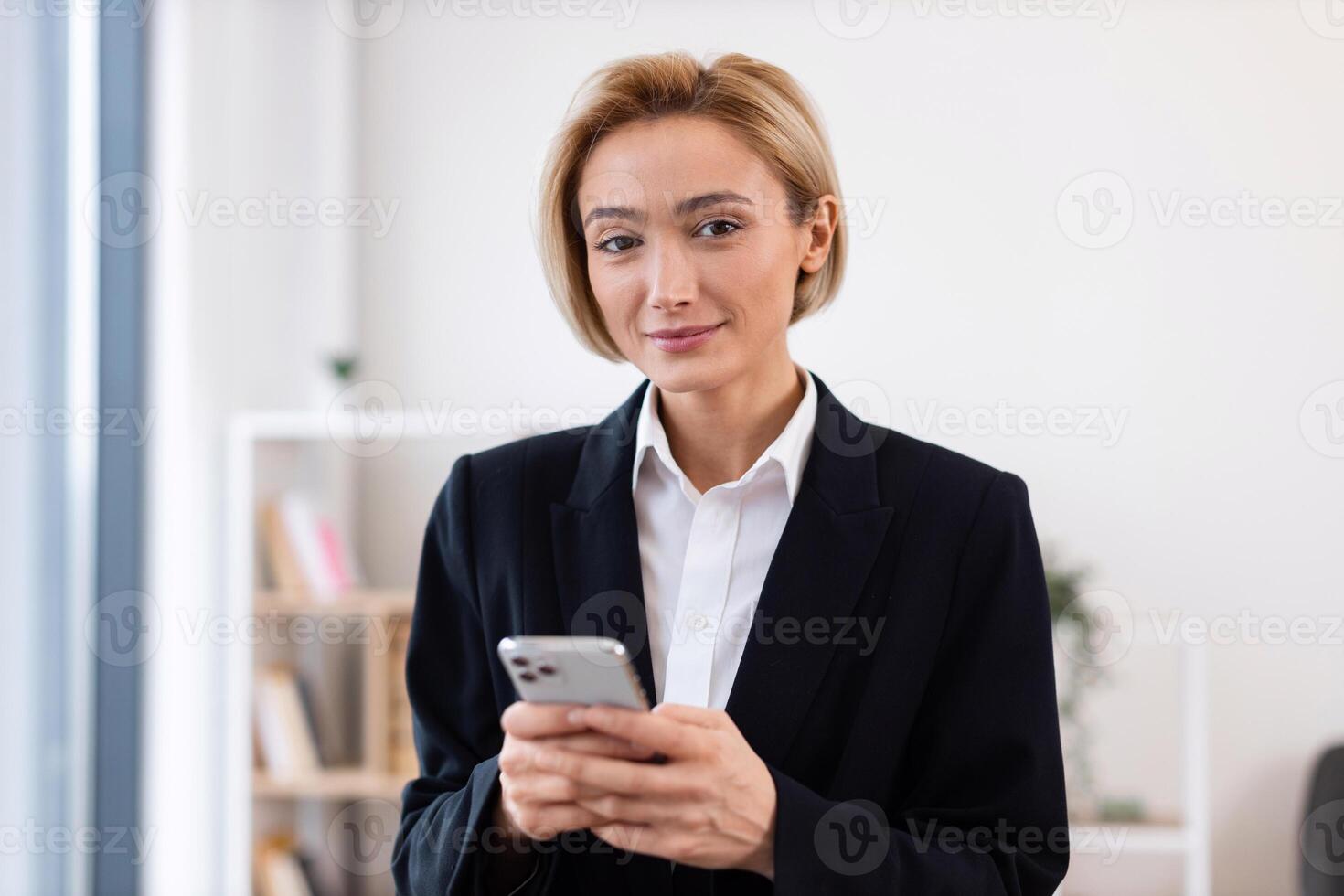 Elegant adult woman wearing formal suit standing indoors holding smartphone looking confident. Represents business communication, technology usage. Professional setting, light and welcoming workspace photo