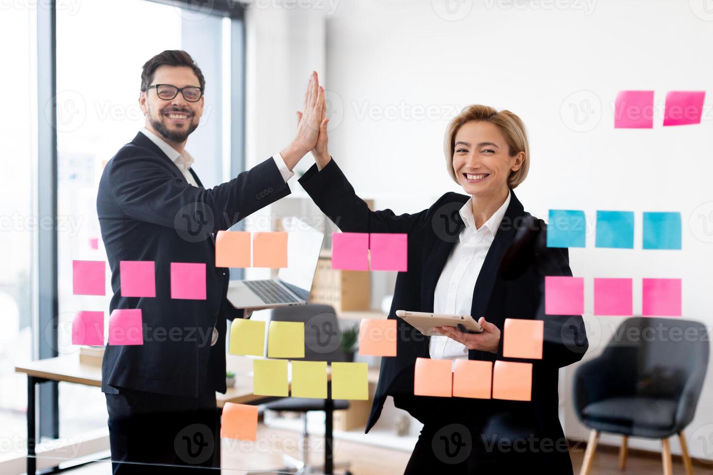 Businessman and businesswoman in formal wear high-fiving, discussing ideas using glass board with sticky notes, cheerful teamwork concept photo