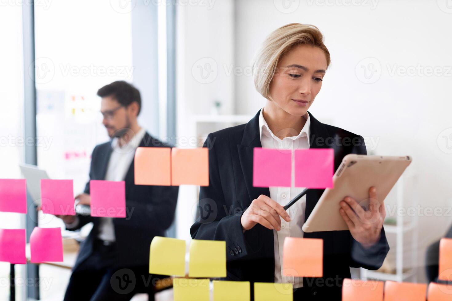 Businesswoman and businessman in professional attire working with sticky notes and devices indoors. Scene represents teamwork, planning, modern business environment, creativity, and productivity. photo
