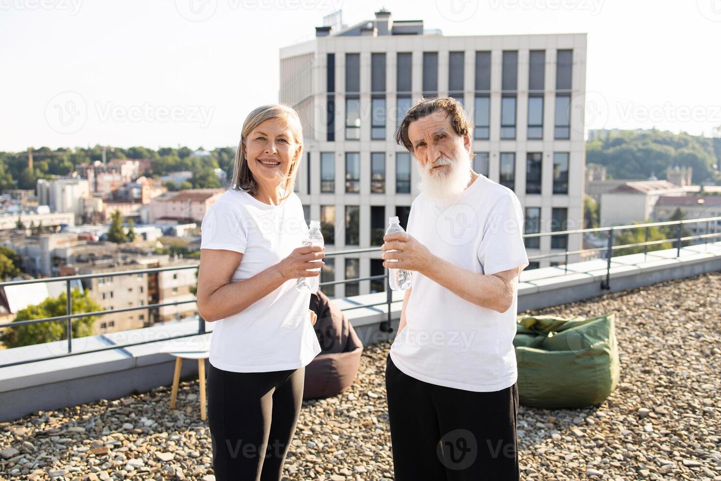 Elderly couple staying hydrated after exercise on urban rooftop during sunny morning. They wear casual sports attire, smile, and enjoy well-being. Cityscape and blue sky provide urban atmosphere. photo