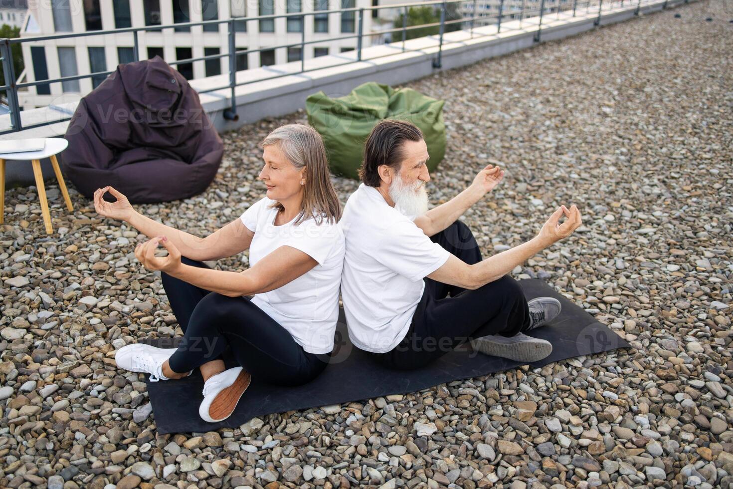 Senior couple performing relaxing meditation seated back to back in lotus pose outdoors. Man and woman wearing white and black activewear. Balanced lifestyle and harmony concept in urban environment. photo