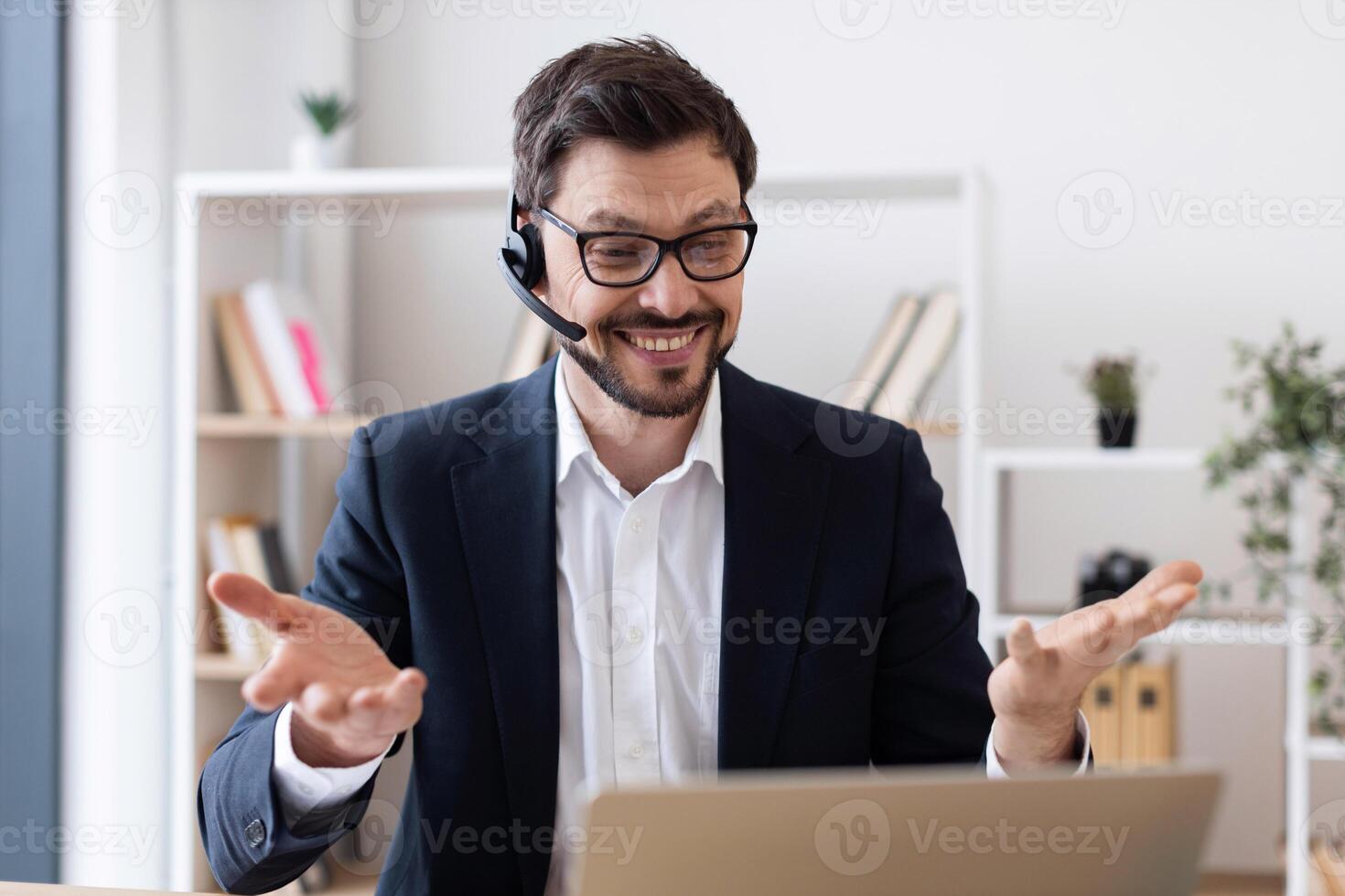 Smiling young man in formal suit using headset and laptop during call in office. Professional working environment with bookshelves in background showcases cheerful approach to remote working. photo