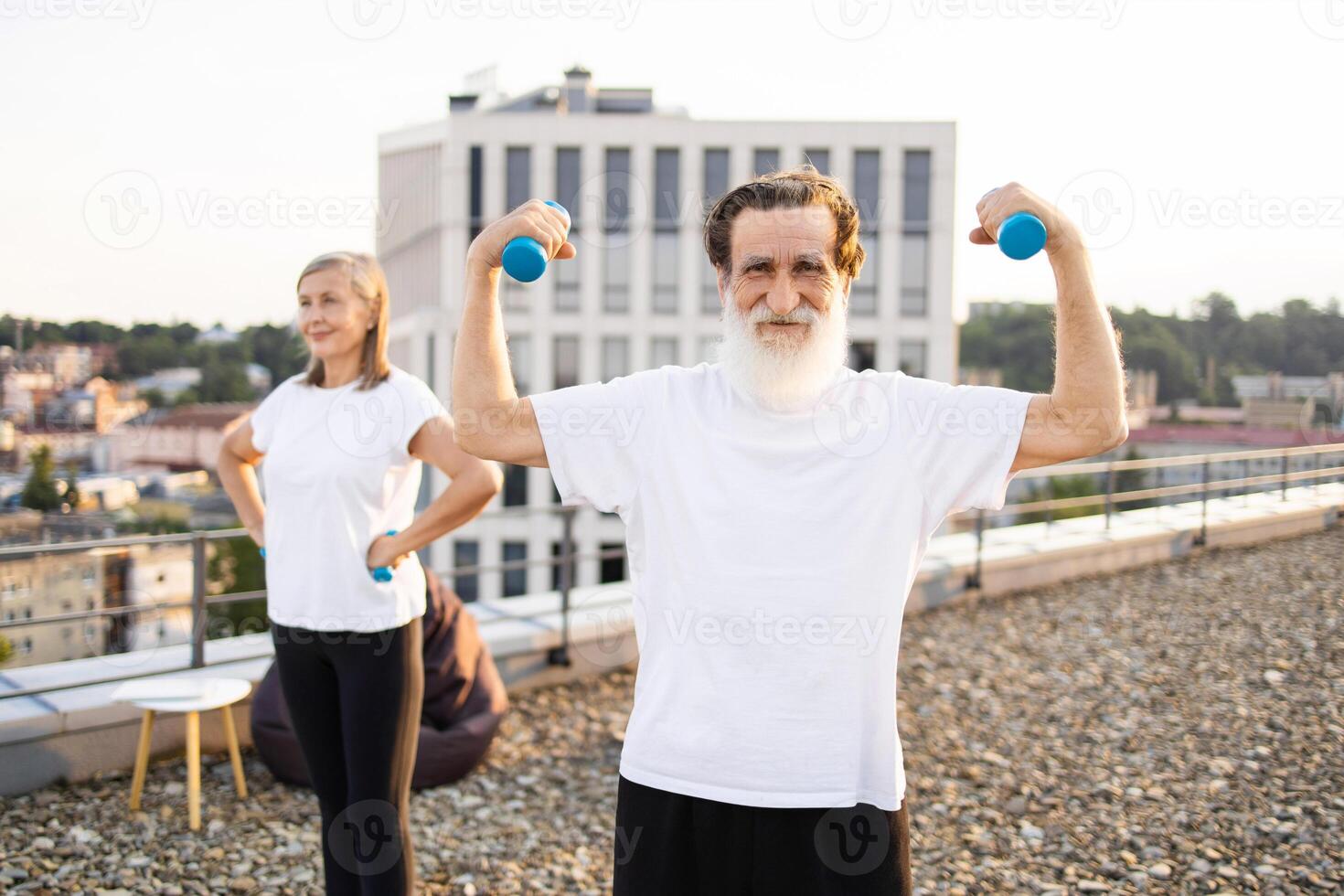 Elderly man and woman outdoors exercising together on rooftop with dumbbells. Healthy lifestyle activity emphasizing fitness, balance, and wellbeing for senior adults in fresh open-air setting. photo