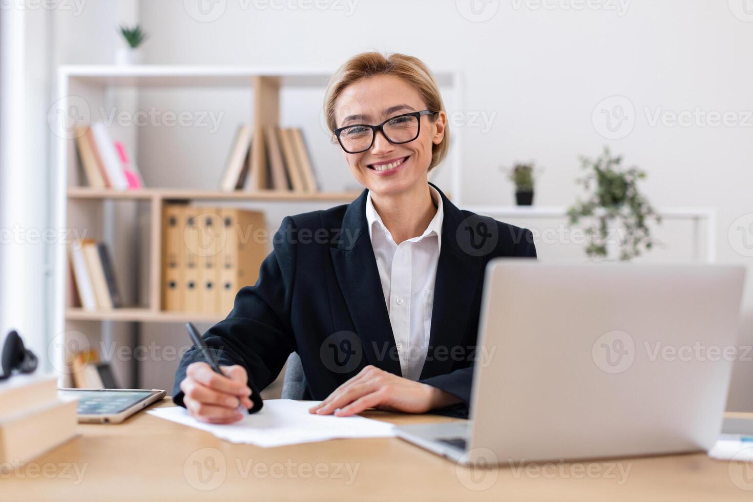 Professional woman wearing business attire writing on paper at desk with laptop in office. Smiling face suggests positivity, productivity. Background shelves hold books plants and organized documents. photo