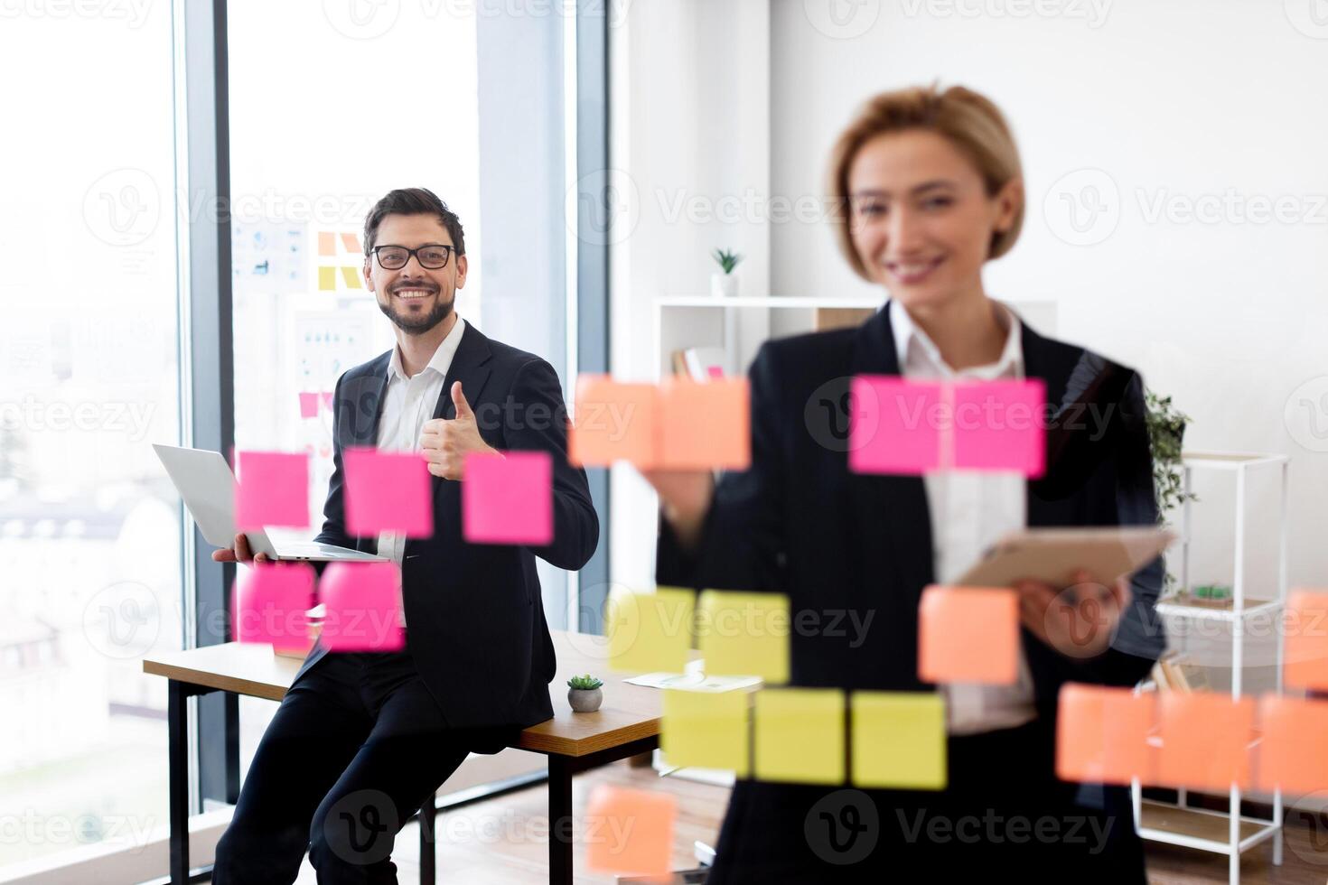 Man and woman in business attire collaborating on project using sticky notes on glass board. Modern office setting with teamwork and productivity focus. photo