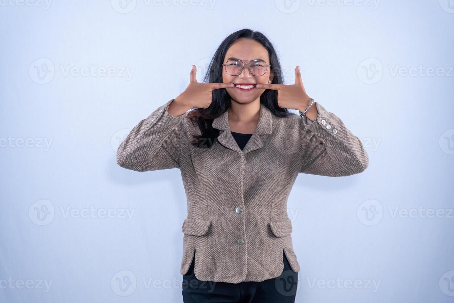 A young woman in a brown jacket and glasses is showing a wide smile with fingers pointing at her cheeks happily. photo
