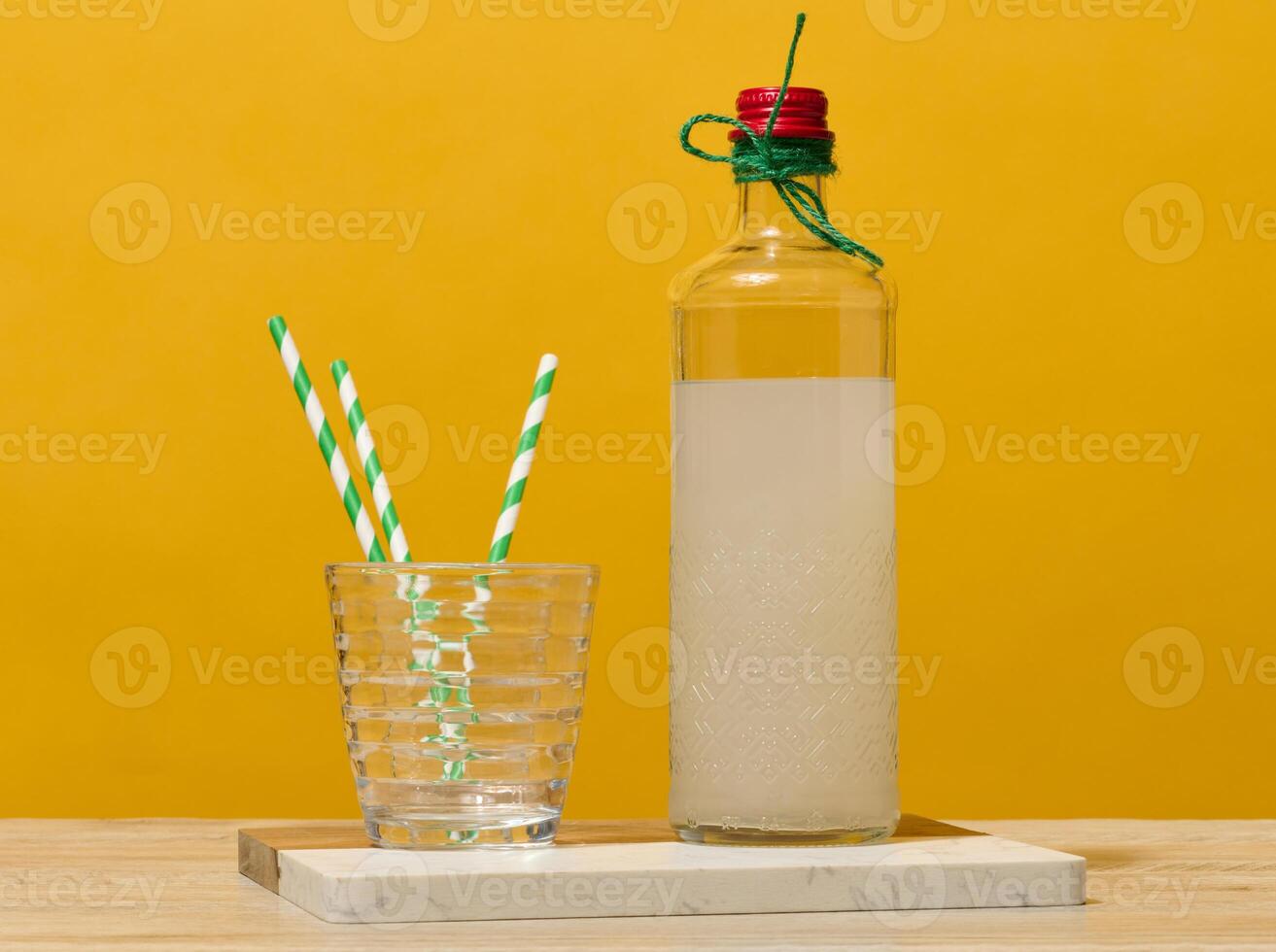 A transparent glass bottle of lemonade stands next to a faceted glass containing three green striped straws. photo