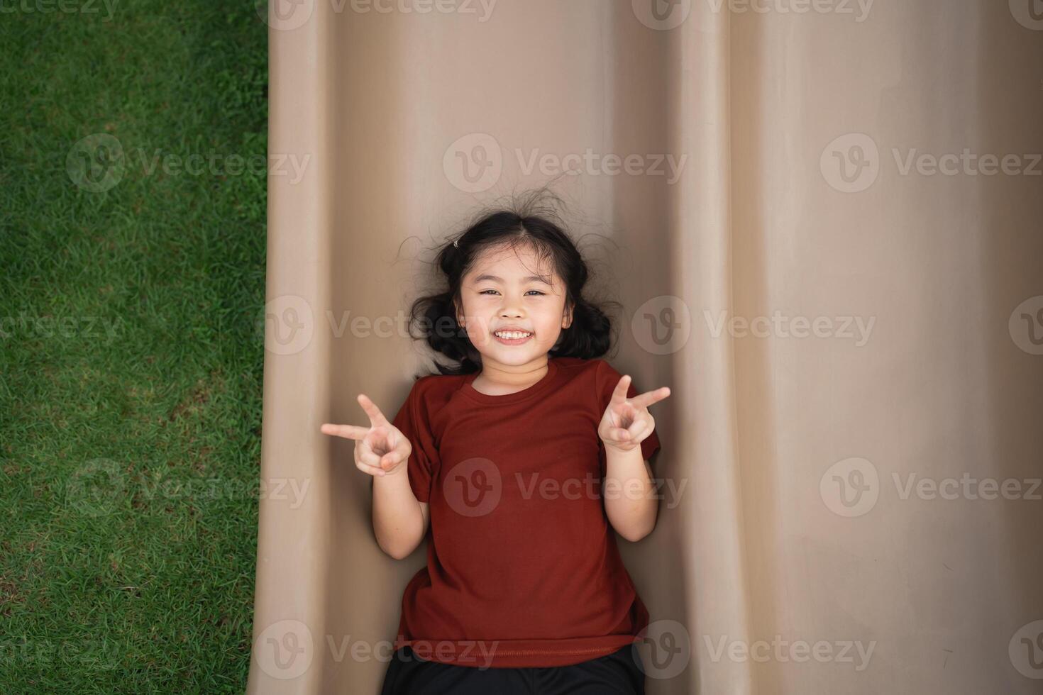 Joyful child playing on a slide outdoors, wearing a brown shirt, smiling while lying on a playground slide with a grassy background photo