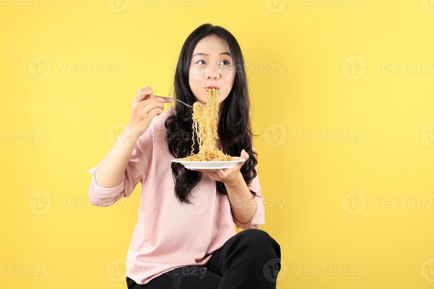 Happy Young Asian Woman Sitting on a Chair Wearing Pink Shirt, Eating Noodles on White Plate using Fork. photo