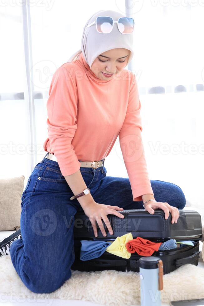 Happy Young Muslim Asian Woman Packing and Trying to Close Luggage. photo
