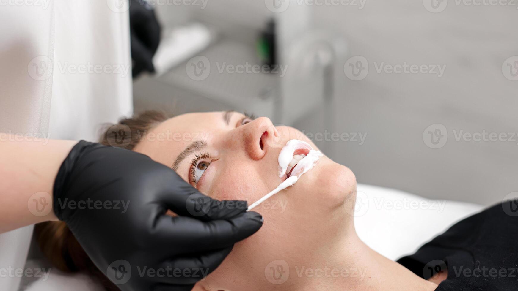 A cosmetologist wearing gloves applies anesthetic cream to a patient's lips before injecting a filler. Concept of cosmetology. photo