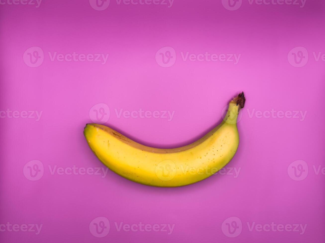 One bright yellow banana lies horizontally on a purple background, top view, centered in the image photo
