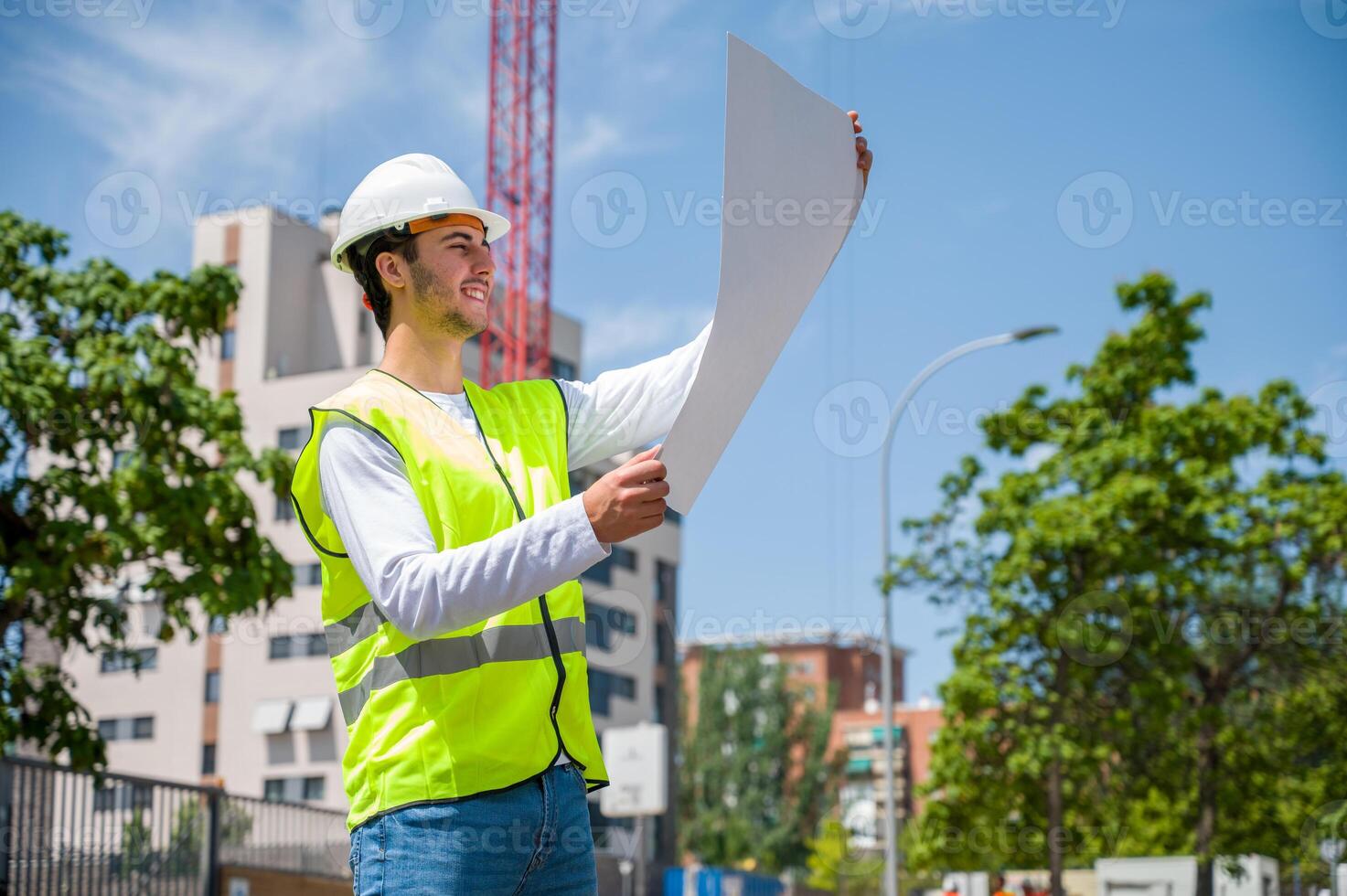 Professional young male construction worker with safety helmet and reflective vest checking work plan at a construction site with building progress in the background. photo