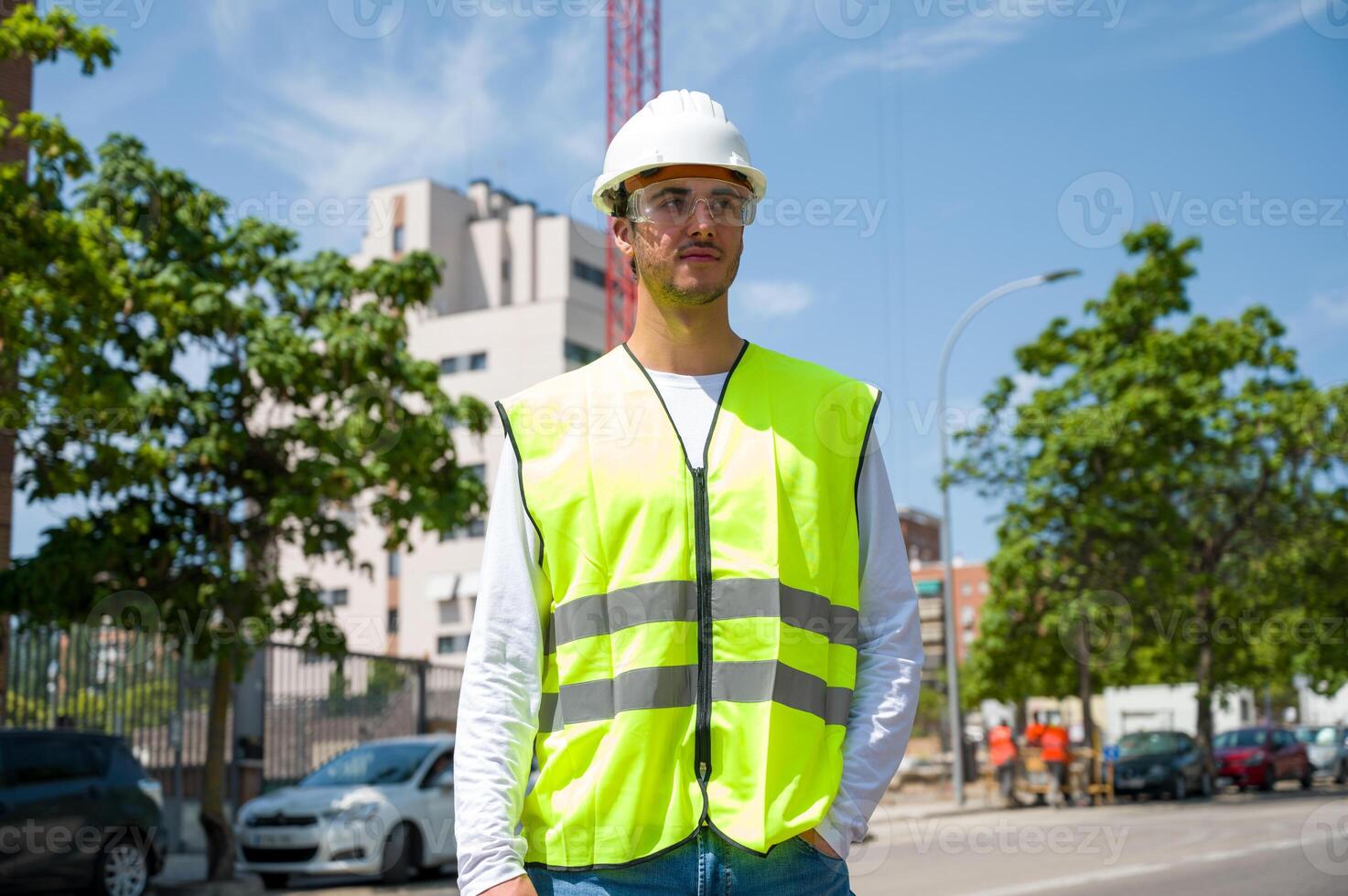 Professional young male construction worker with safety helmet and reflective vest at a construction site with building progress in the background... photo