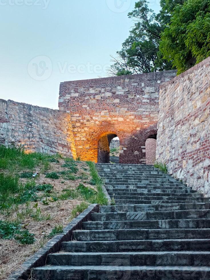 Stone steps lead to an illuminated archway in an old fortress. The path is flanked by historic brick and stone walls, leading the viewer into a serene twilight setting. photo