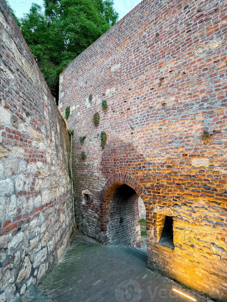 Narrow medieval alley framed by ancient brick walls, featuring an arched tunnel and small window niche. Soft lighting enhances the aged texture of this fortress corridor photo