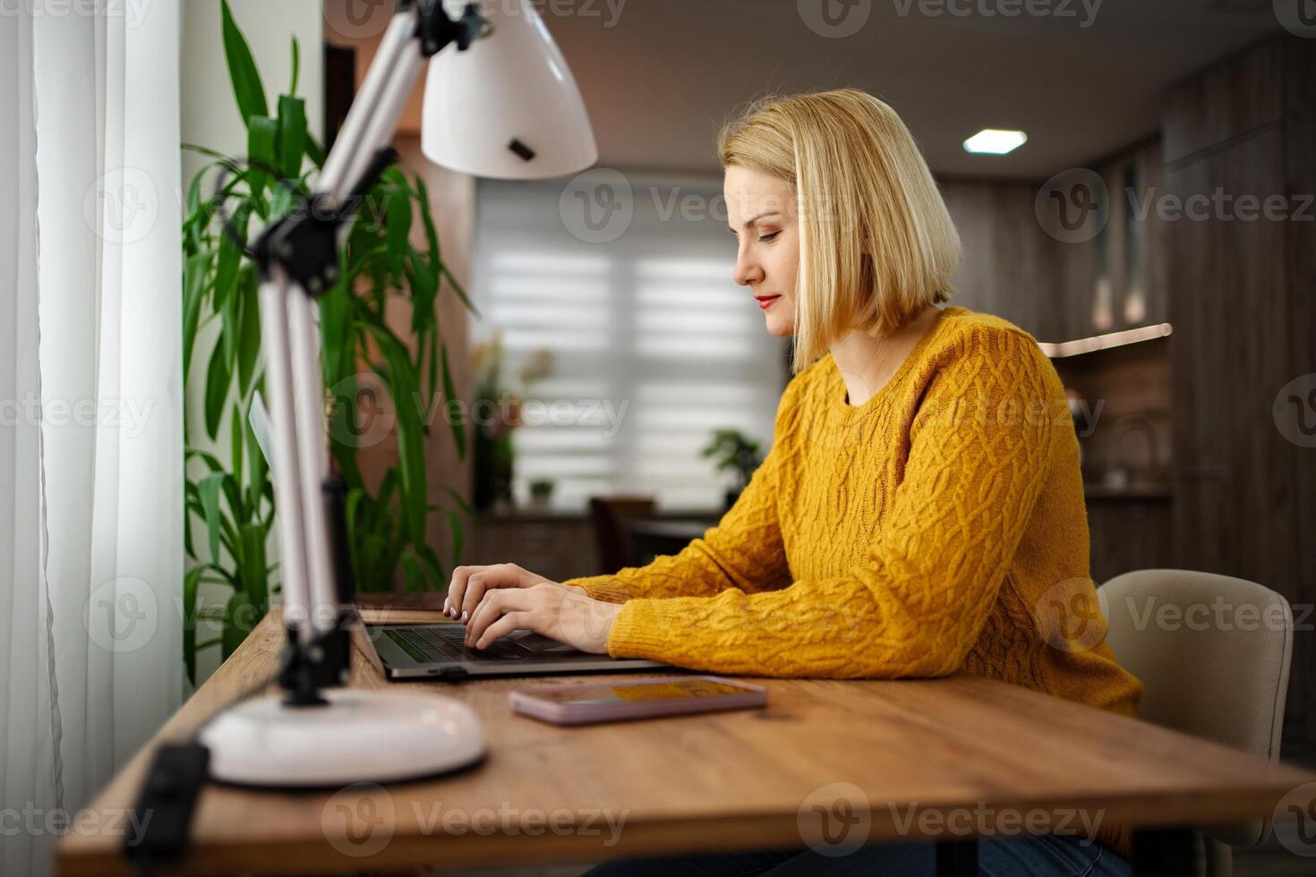 Focused woman in yellow sweater working on laptop at home desk with bright lamp and plants in a cozy atmosphere photo