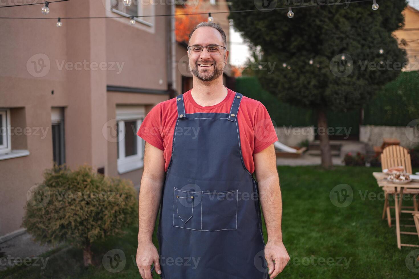 Smiling man in apron stands in a vibrant backyard during sunset gathering photo