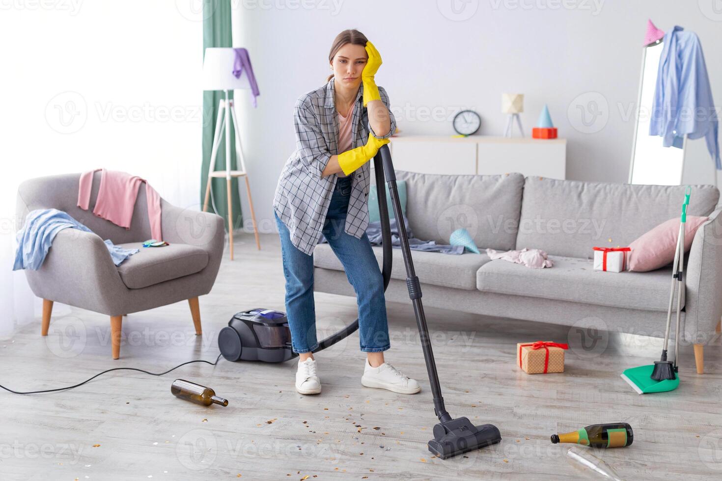 Unhappy young woman standing with vacuum cleaner in messy flat after party, blank space. Upset millennial housewife at dirty room after celebration, garbage scattered everywhere photo