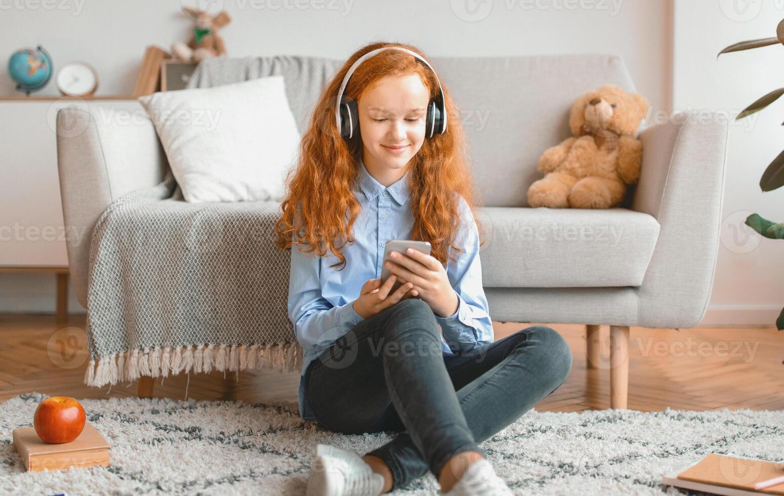 Leisure Concept. Smiling Red-Haired Schoolgirl Sitting On The Floor Leaning On The Couch At Home, Listening To Music In Wireless Headphones And Chatting On Her Cell Phone, Enjoying Free Time photo