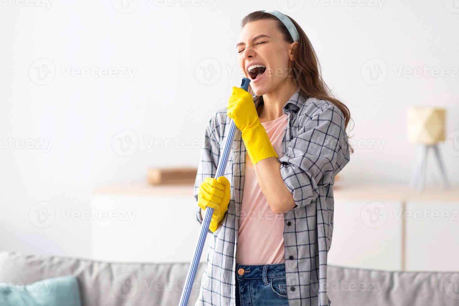 House cleaning is fun. Cheerful young lady using mop as mic, singing song while doing cleanup at living room. Millennial housewife pretending to be rock star while doing domestic duties photo
