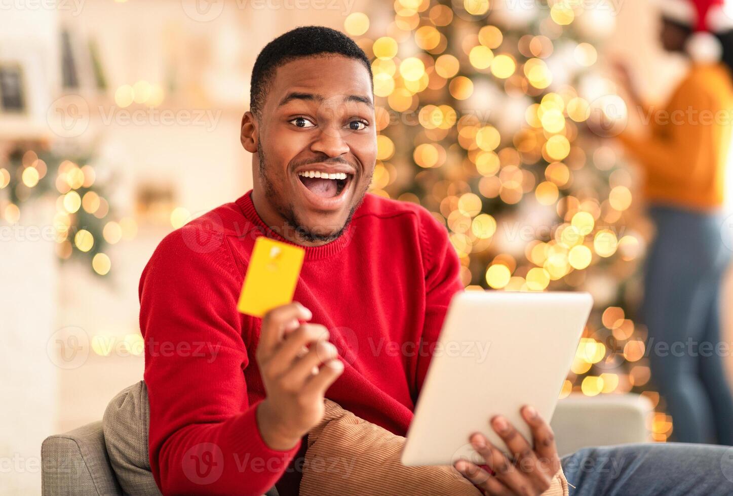 Super offer from store, cashback and online shopping. Successful millennial african american husband holds tablet and credit card in interior of living room, woman in santa hat decorating Xmas tree photo