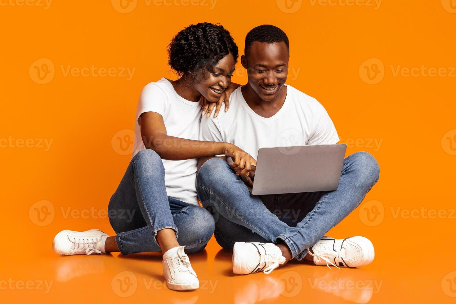 Relaxed african american man and woman using new laptop together while sitting on floor, checking social media, shopping online or watching photos, orange studio background, copy space photo